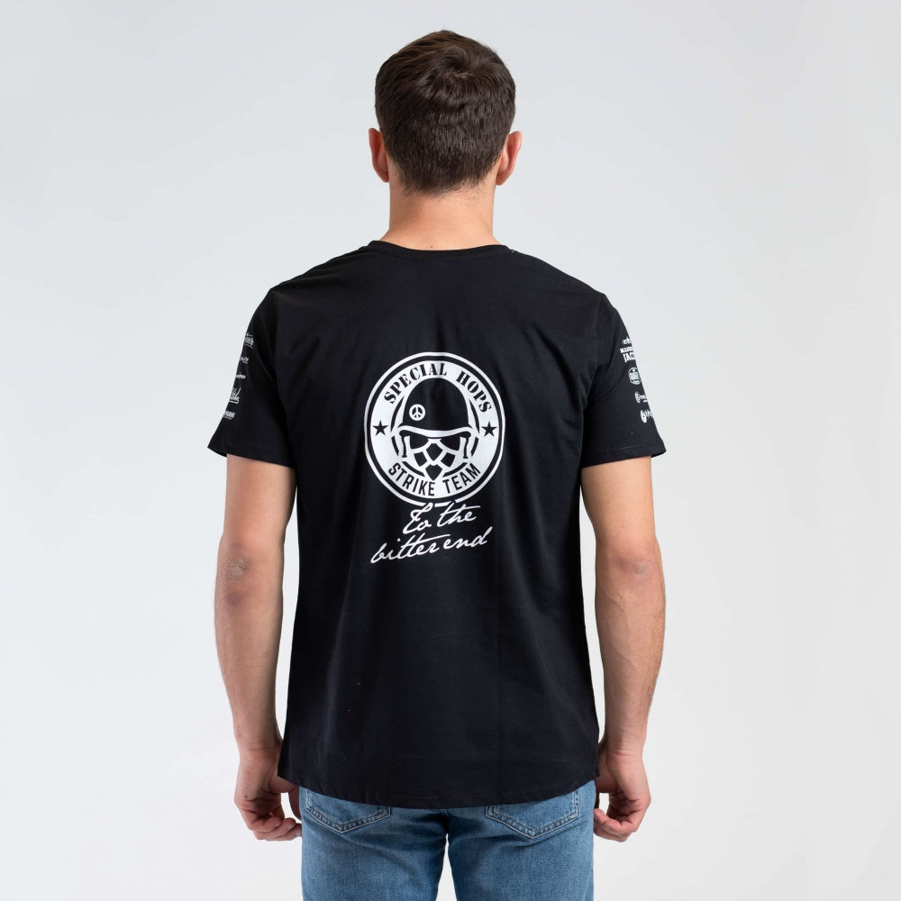 En mann med ansiktet vendt mot seg, iført Brewshop Craft Brewers Team T-skjorte - unisex, med en sirkulær Special Move Strike Team-grafikk og tekst på ryggen, sammen med blå jeans.