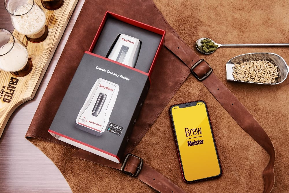 Et Anton Paar EasyDens Digitalt Hydrometer i esken, en telefon med Brew Meister-appen, malt bygg, humle, skinnforkle og ølglass på et Crafted-brett - ideelt for øl- og vinfans - plassert på en brun flate.
