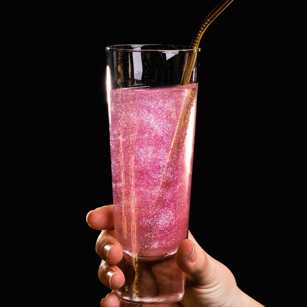 En hånd holder et høyt glass med rosa, glitrende drikke laget med Eco Lovers Eat Me Glitter Rose 20 ml og et sugerør i gull, mot en svart bakgrunn.