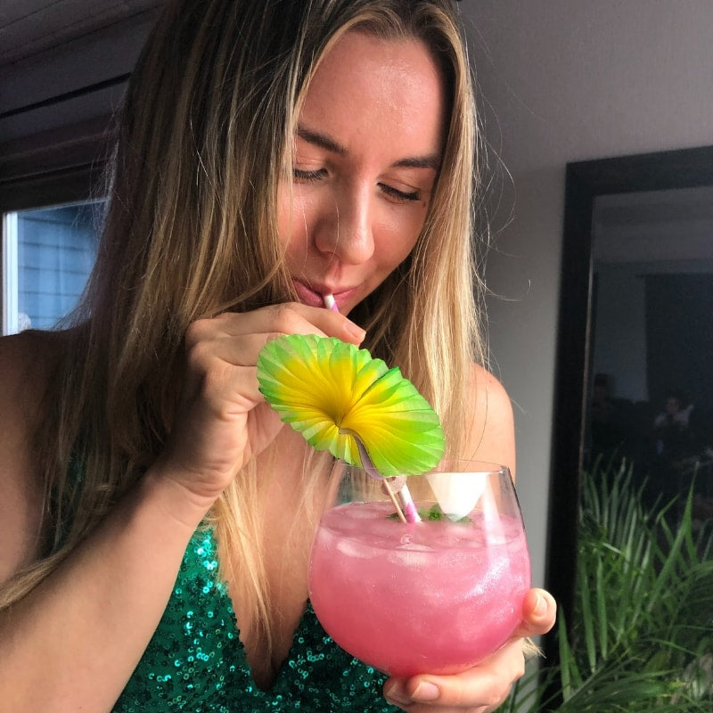 En kvinne i grønn paljettoverdel nipper til en rosa cocktail laget med Eco Lovers Eat Me Glitter Rose 20ml, servert på is med en dekorativ paraply og sugerør.