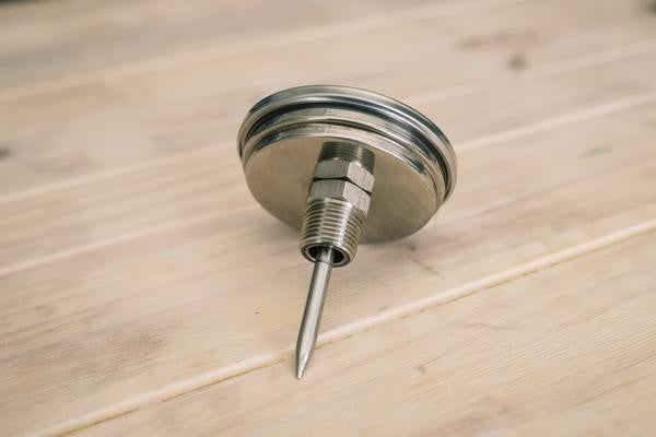 A Termometer for Ss Brew Kettle m/gjennomføring fra Ss Brewtech med metallsonde og 20mm gjenger i rustfritt stål ligger på en treoverflate.
