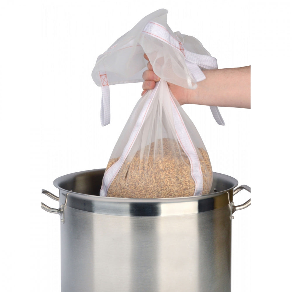 En hånd løfter The Brew Bag 50L - 40 x 47cm Meskepose fra Brew in a bag supply, fylt med korn, ut av en stor bryggekjele, klar for neste bryggetrinn.