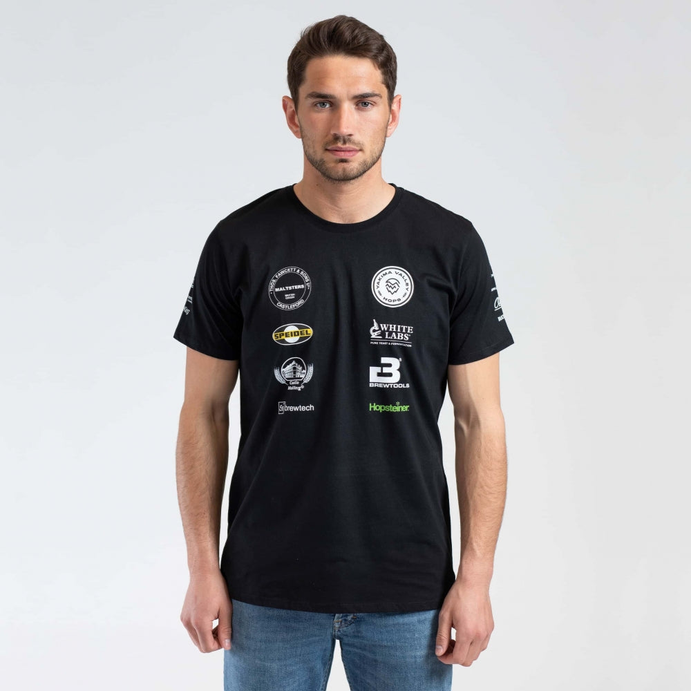 En mann har på seg Brewshop Craft Brewers Team T-skjorte - Unisex, med flere sponsorlogoer på forsiden, mot en vanlig lys bakgrunn.