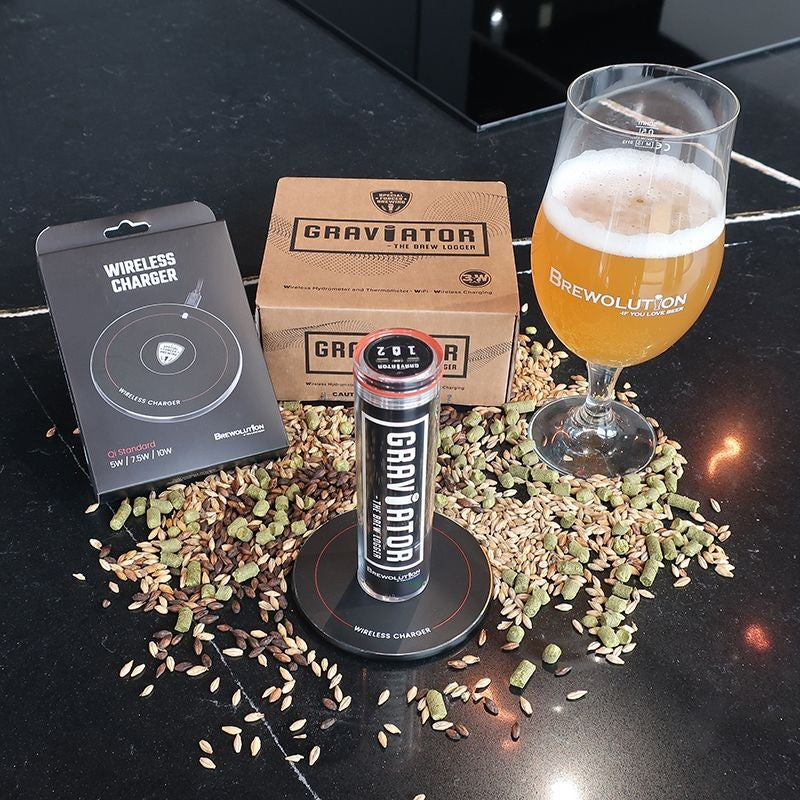En Brewolution Graviator - WiFi-hydrometer og termometer, emballasje og et ølglass står på en benk overstrødd med sølt korn og kardemommekapsler.
