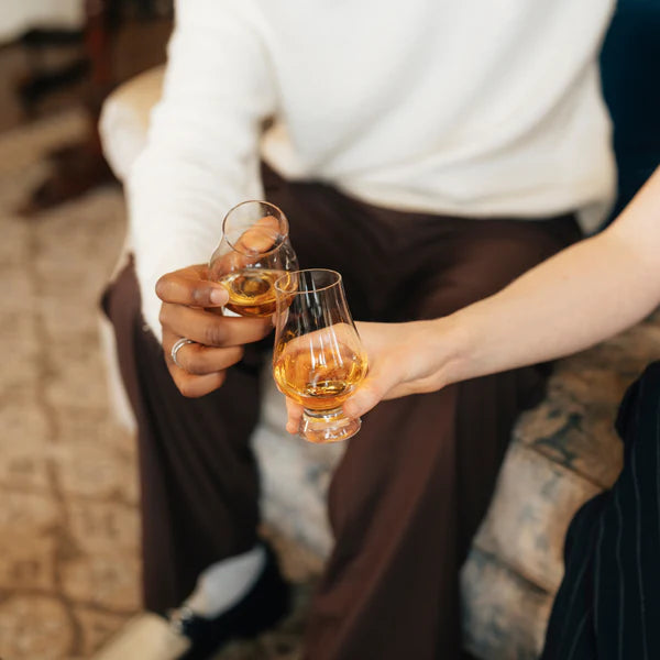 To personer skåler i Glencairn Whisky Glass 190 ml fra Stölzle Lausits, hver fylt med ravfarget brennevin, mens de slapper av i en sofa.