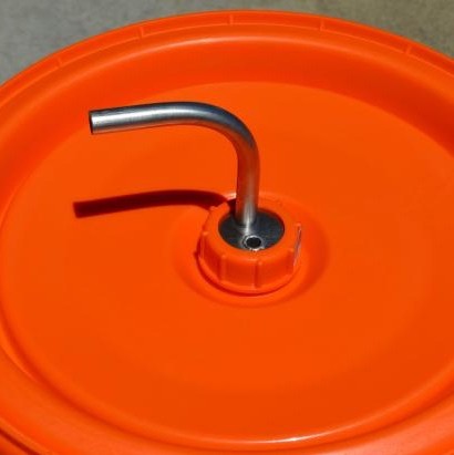 Nærbilde av Speidel Fermenter Blow Off Tube Thermowell - Norcal (Termobrønn): et oransje plastlokk med en 304 rustfri stålstuss fra Norcal, festet gjennom midten for bruk som et avblåsningsrør eller termohull.