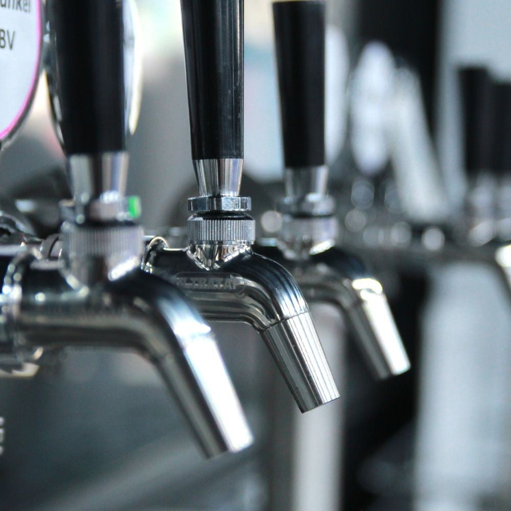 Nærbilde av flere ølkraner i metall med svarte håndtak, blant annet en Keg King Ultratap SS tappekran - med håndtak i rustfritt stål, oppstilt på rekke og rad i en bar eller et bryggeri.
