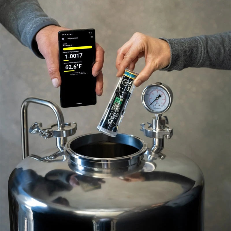 To personer bruker Tilt Hydrometer Tilt Pro Hydrometer & Termometer og app for å sjekke temperaturen i en gjæringstank av rustfritt stål ved siden av en analog måler, samtidig som de følger med på stabiliteten i den spesifikke vekten.