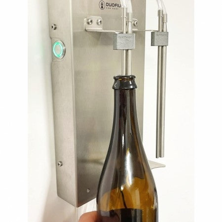 En hånd holder en brun glassflaske under Duofiller Core (G3) Automatic Can Filler fra Duofiller, en enhet i rustfritt stål som er montert på veggen - et ideelt oppsett for enhver hjemmebrygger.