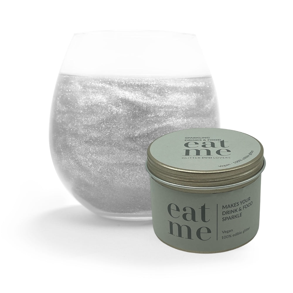 Et glass med en glitrende drink sitter bak en Eco Lovers Eat Me Glitter Clear 150 ml beholder, fylt med spiselig glitter - perfekt for å gi enhver godbit eller drikke et blendende skimmer.