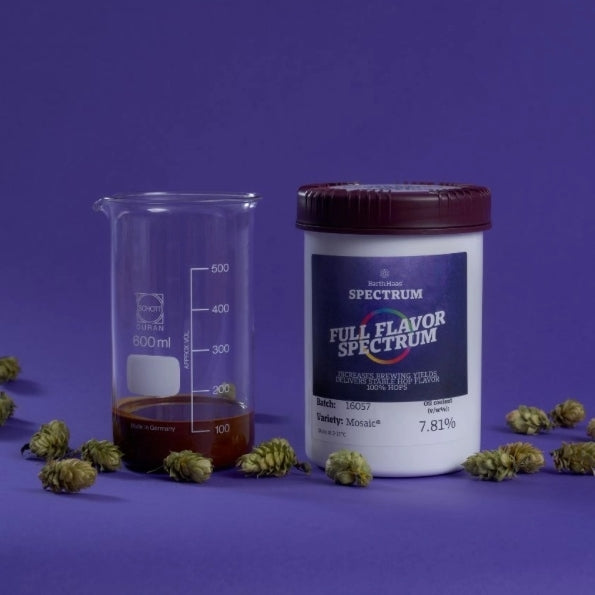 Et beger med ravfarget væske og en Cascade Spectrum 1 kg (Best før 19.12.25) står på en lilla flate omgitt av humlekjegler som fremhever den livlige aromaen i denne flytende humlen.