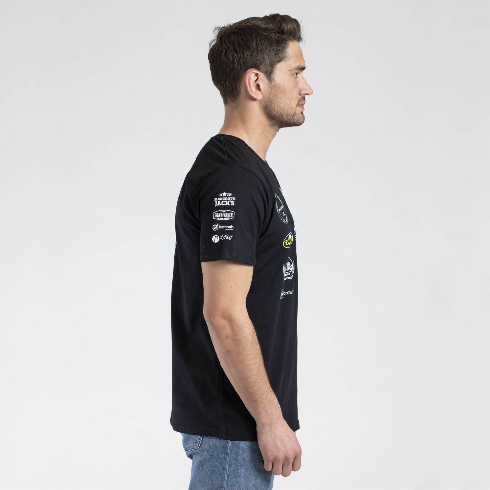 En mann står i profil med ansiktet vendt mot høyre, iført en svart Brewshop Craft Brewers Team T-Shirt - Unisex med hvit logo og blå jeans mot en vanlig lys bakgrunn.