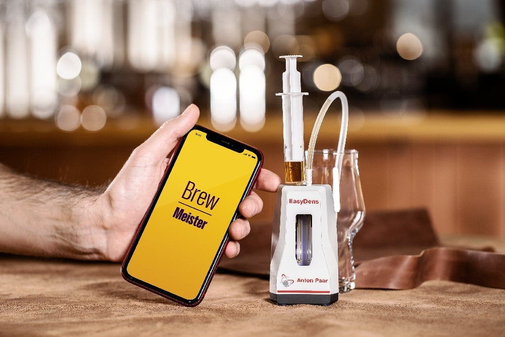 En hånd holder en smarttelefon som viser "Brew Meister" ved siden av Anton Paar EasyDens Digitalt Hydrometer og en sprøyte på et bord med et tomt glass - ideelt for øl- og vinentusiaster.