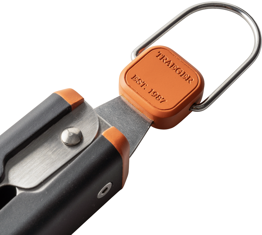 Nærbilde av Traeger BBQ Tongs by Traeger - svart og sølv grillklype med oransje aksenter, metallsløyfe og "TRAEGER EST. 1987"; uunnværlig BBQ tilbehør laget av rustfritt stål for grilling.