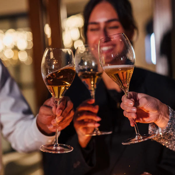 Tre personer skåler i Stölzle Lausits Fino champagneglass 425 ml fra et 6-delt sett, fylt med champagne, mens en person smiler i bakgrunnen.