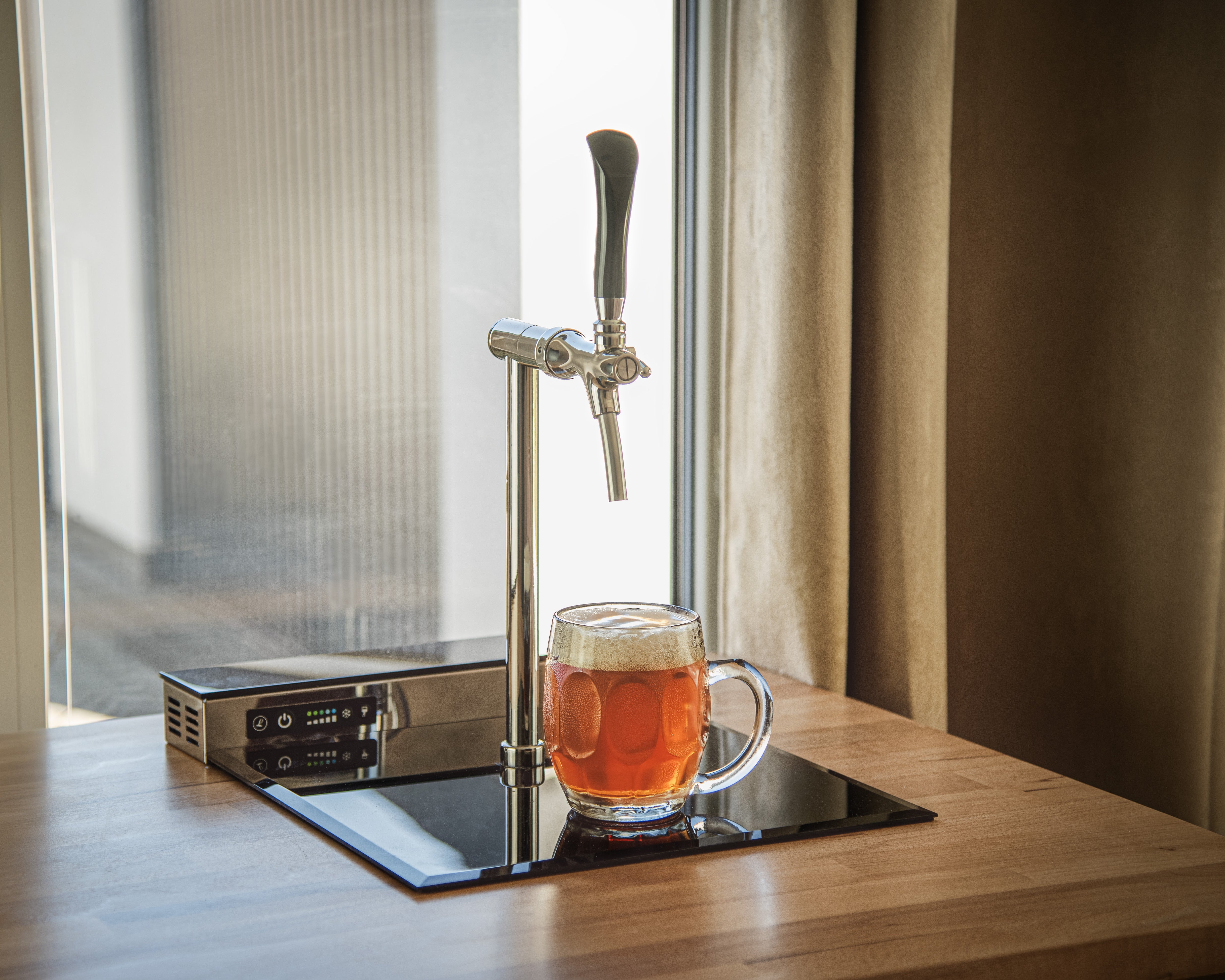 Et Lindr PYGMY Pro Kitchen øltappesystem fra Lindr skjenker øl i et glasskrus på en benkeplate av tre i nærheten av et vindu med beige gardiner.