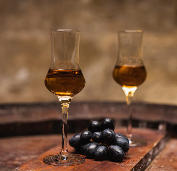 To glass dessertvin serveres i et elegant Stölzle Lausits Grappaglass 90 ml, som hviler på en treflate ved siden av en liten klase svarte druer.