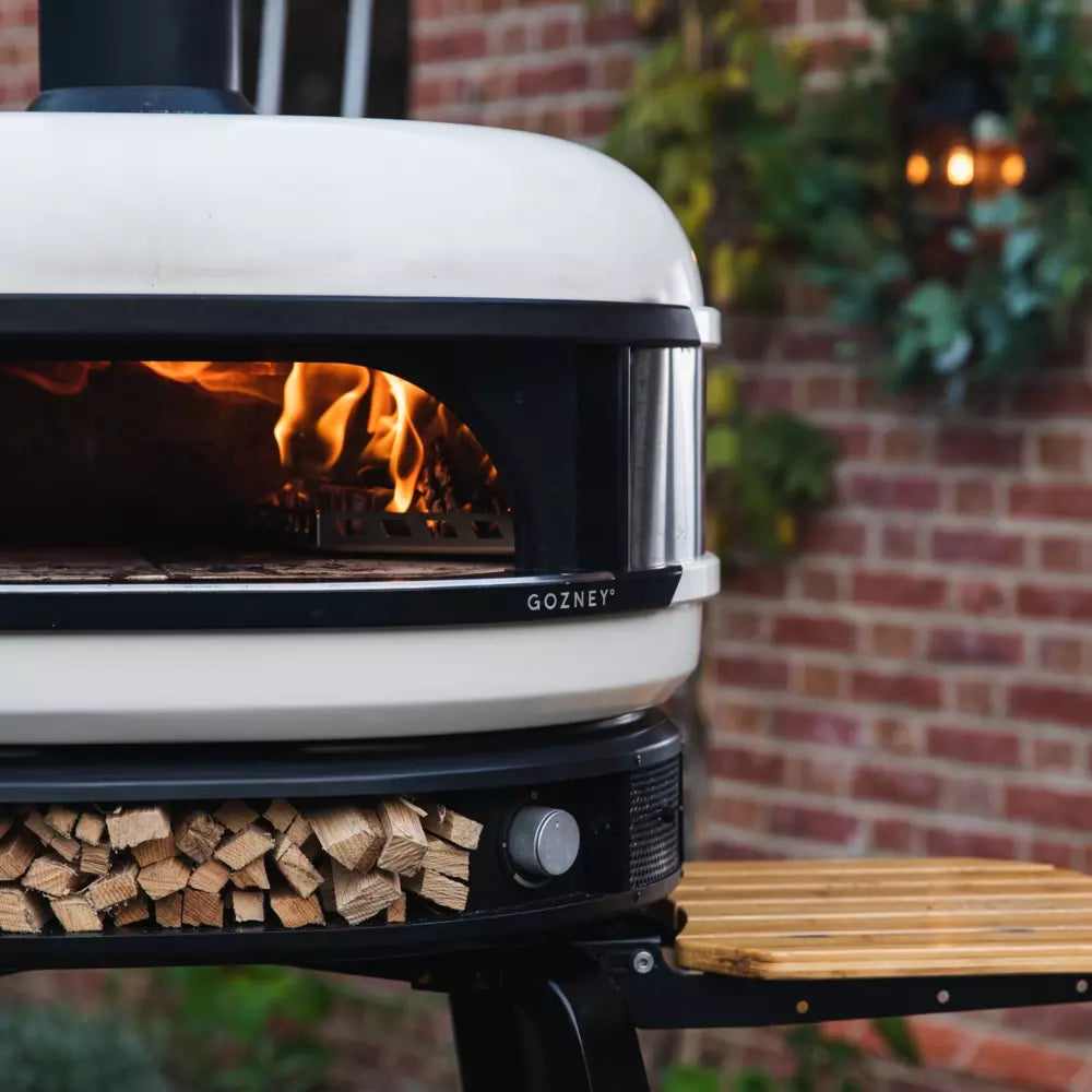 Et nærbilde av Gozney Dome - Bone - Multi Fuel Pizzaovn fra Gozney med et bål som brenner på innsiden og stablet ved nedenfor, omgitt av en murvegg - perfekt for baking av autentisk napolitansk pizza.