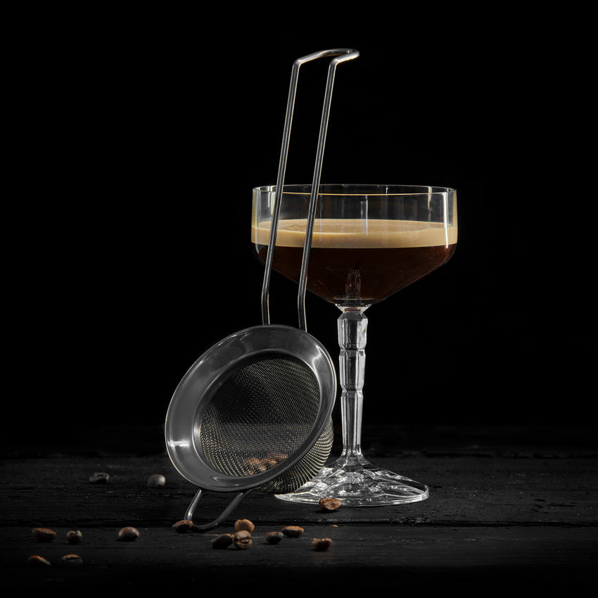 Et coupeglass med espresso martini står bak Lurch Fin Cocktailsil, med kaffebønner spredt utover en mørk overflate, og viser frem elegante barredskaper for den perfekte cocktailopplevelsen.