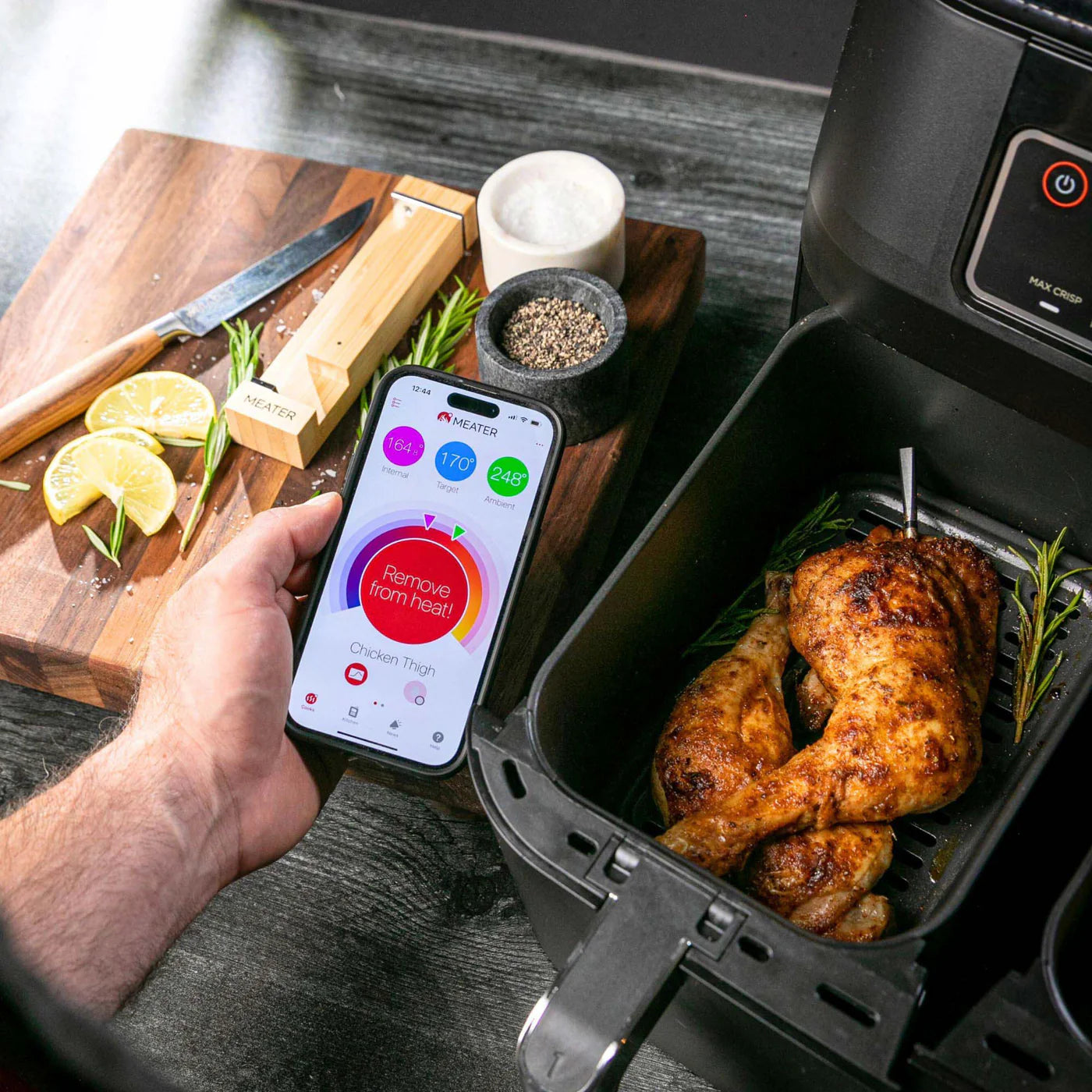 En hånd holder en smarttelefon med Traeger Meater Pro - Bluetooth-steketermometer-appen ved siden av en frityrgryte med stekte kyllinglår; like ved står et skjærebrett med krydder og sitron.