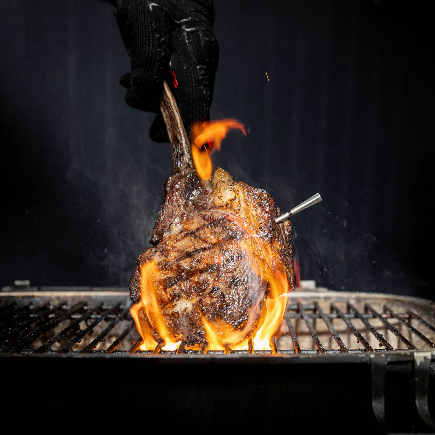En hanskekledd hånd holder en grillet biff med et Traeger Meater Pro - Bluetooth steketermometer mens flammene står opp fra grillen under.