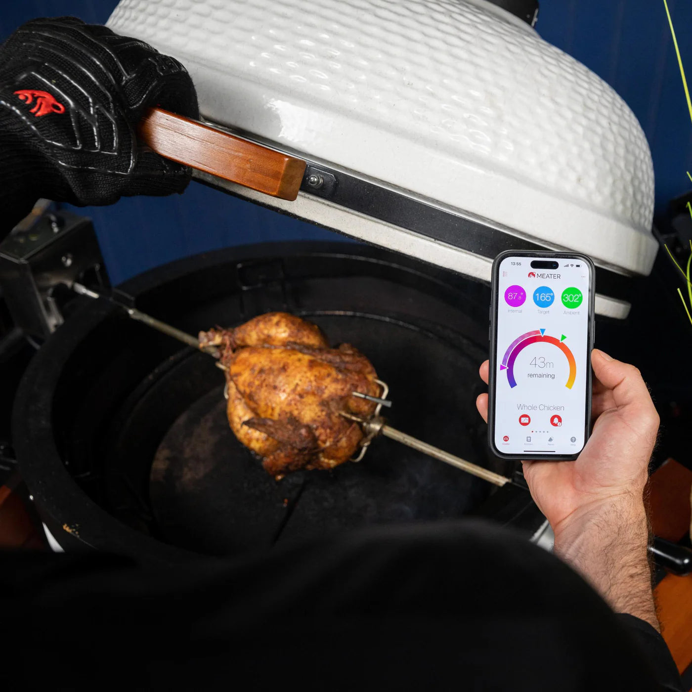 En person bruker Traeger Meater Pro - Bluetooth steketermometer-appen på smarttelefonen sin for å sjekke timeren og temperaturen mens han eller hun overvåker en rotisseriekylling som stekes i en keramisk grill.