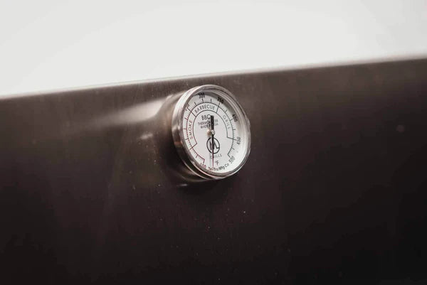 Et nærbilde av en M Grills B24 Stainless Steel Built In Charcoal Grill viser termometeret rustfritt stål på en mørk overflate, og viser temperaturavlesninger i både Fahrenheit og Celsius.