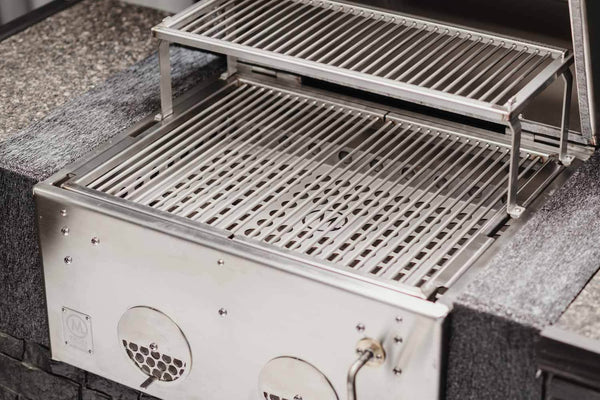 Nærbilde av M Grills B24 Built In Charcoal Grill i rustfritt stål, med to grillhyller og en justerbar kullrist, plassert i en benkeplate av stein.