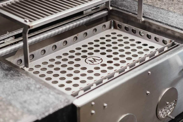 Nærbilde av M Grills B24 Built In Charcoal Grill i rustfritt stål, med perforert brett, justerbar kullrist, hevet varmestativ og en sirkulær M-logo i midten av brettet.