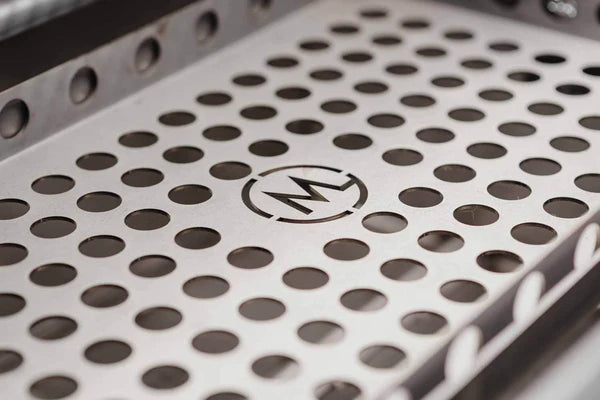 Nærbilde av B24 Stainless Steel Built In Charcoal Grill-skuffen fra M Grills, med metallfinish, jevnt fordelte runde hull og en M-logo i midten - perfekt for grilling med denne førsteklasses rustfritt stål kullgrillen.