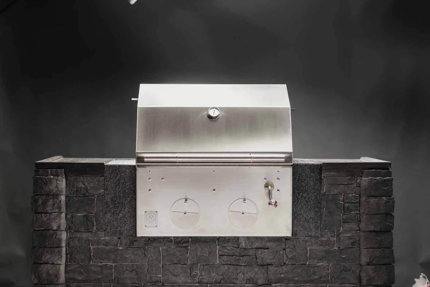 B24 Stainless Steel Built In Charcoal Grill fra M Grills er innebygd i en mørk steinbenk i utekjøkkenet, med lukket lokk og justerbar kullrist mot en svart bakgrunn.