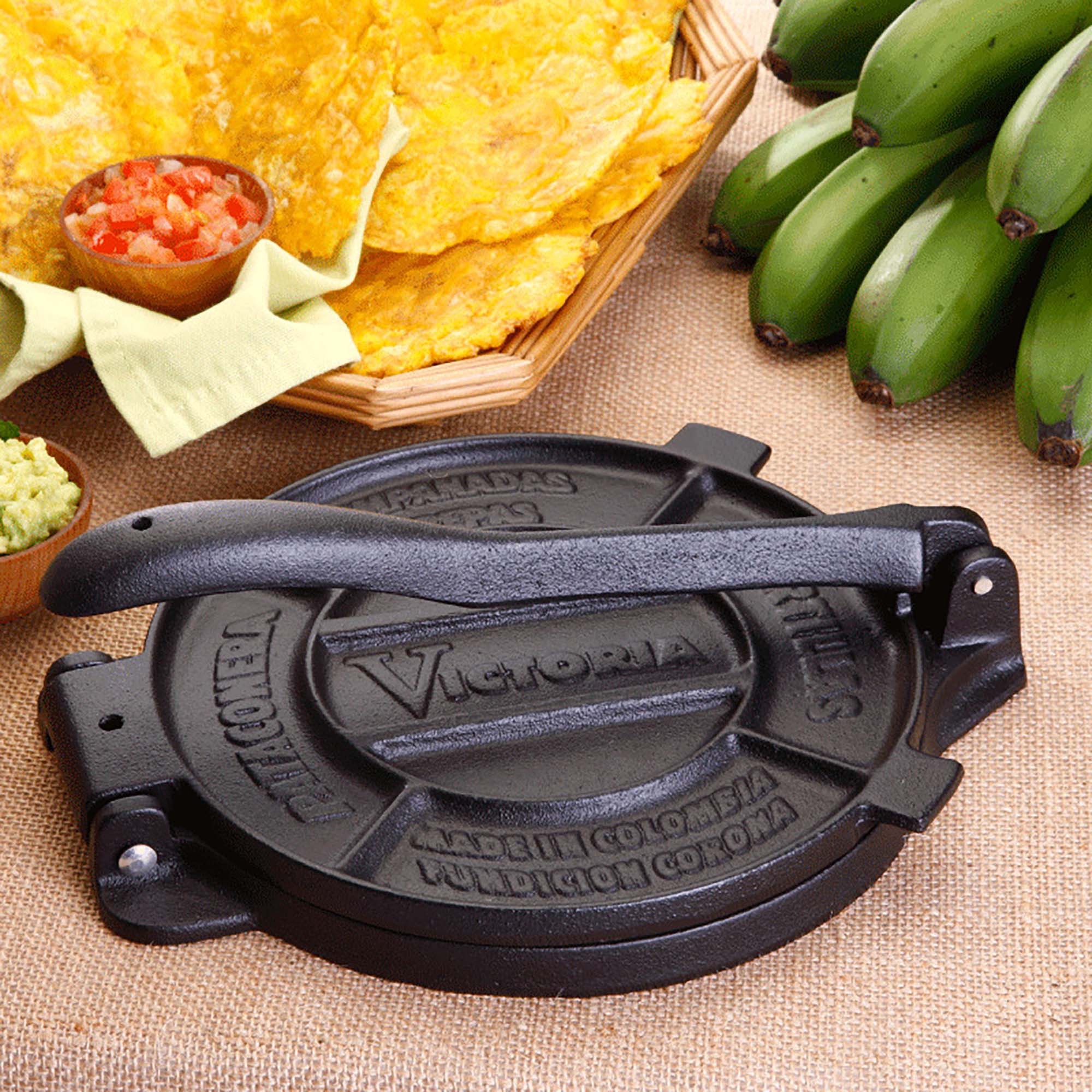 Victoria Tortillapresse 16 cm i Støpejern står på en sækkelærred, omgitt av grønne kokebananer, guacamole, salsa og stekte kokebanankaker - perfekt for å lage hjemmelagde tortillas eller maistortillas hjemme.