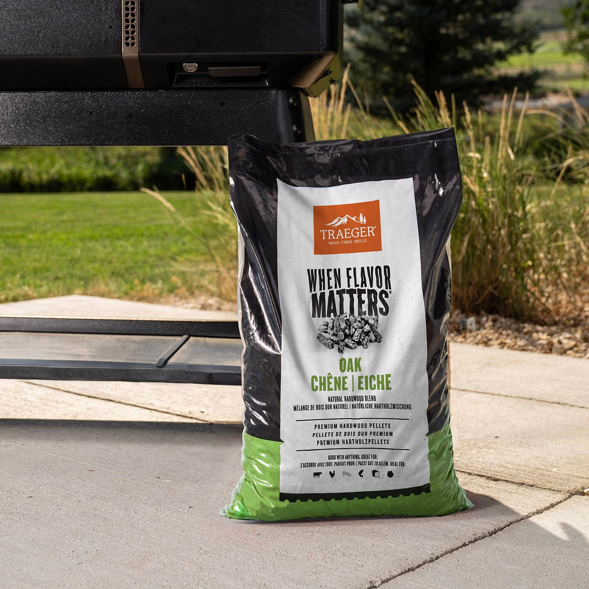 En pose Traeger Oak Pellets 9 kg fra Traeger ligger på en uteplass ved en svart grill, klar til å gi grillen din autentisk røyksmak, med gress og trær i bakgrunnen.