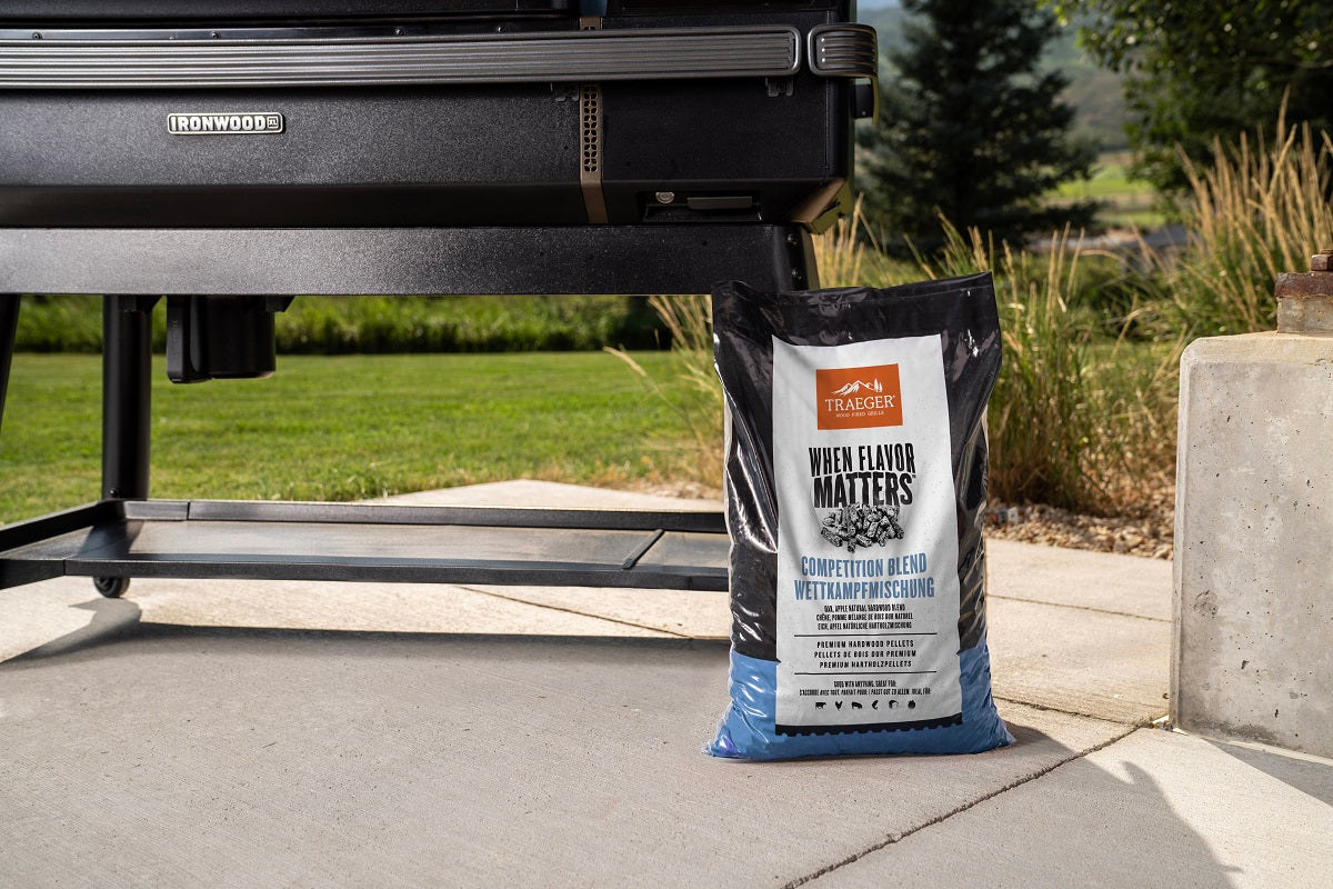 En 9 kg tung sekk med Traeger Competition Blend Pellets står på terrassen ved siden av en svart Ironwood-grill, som leverer 100 % trevirke for ekte røyksmak, med gress og trær i bakgrunnen.