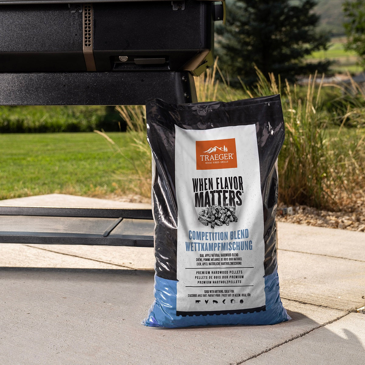 En 9 kg tung sekk med Traeger Competition Blend Pellets fra Traeger ligger på terrassen ved grillen, klar til å tilføre maten rik vedfyrt røyk, med gress og trær i bakgrunnen.
