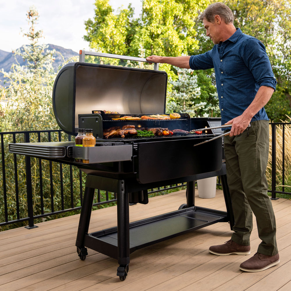 En mann griller diverse matvarer på en Traeger Ironwood XL pelletsgrill med lokket åpent, mens han står på et tredekke mellom trær og fjell og nyter den rike vedfyrt-smaken av matlaging utendørs.