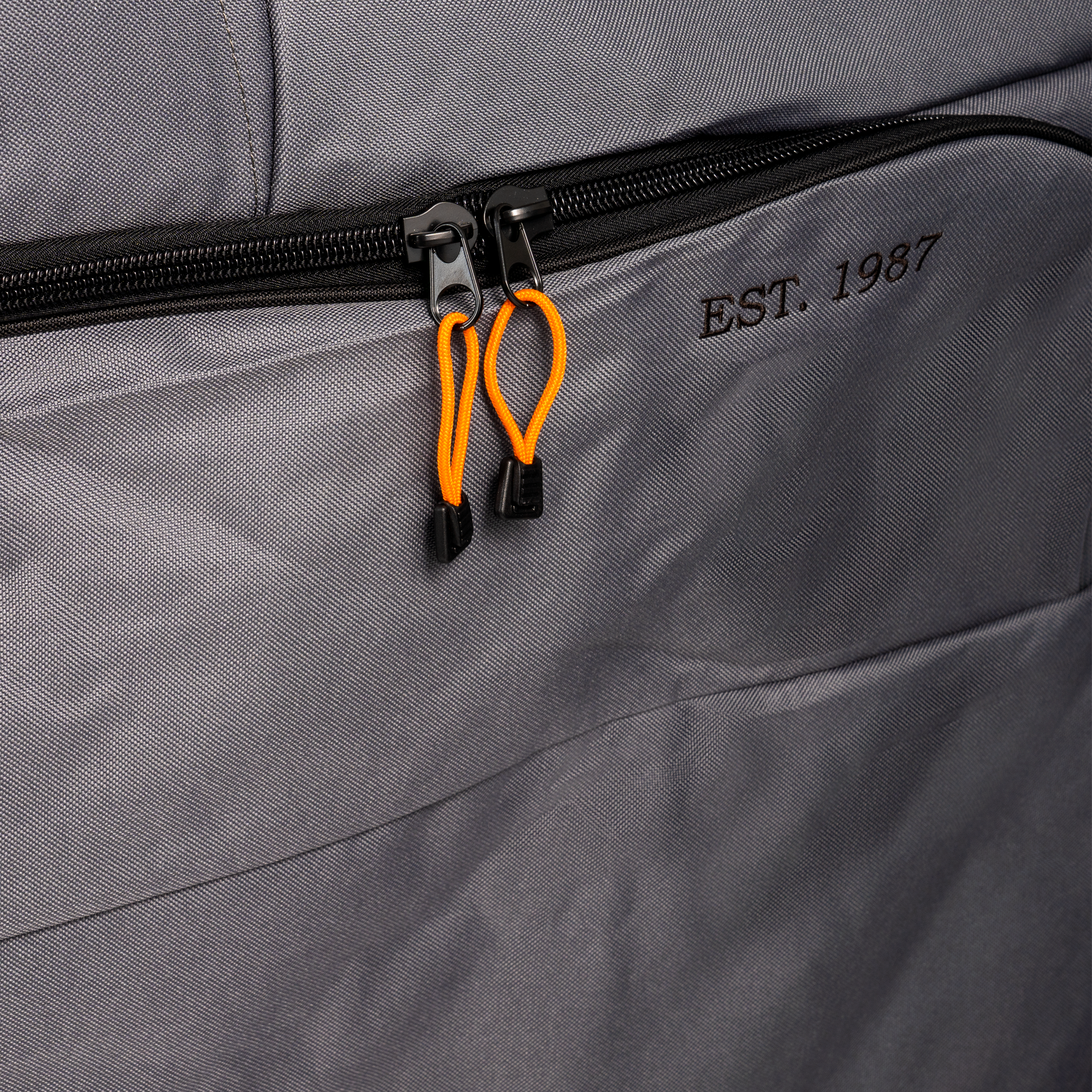Nærbilde av en grå stoffpose med glidelås med to oransje trekkfliker og "EST. 1987" brodert, laget av et materiale som ligner på Traeger Full Length Grill Cover for Timberline XL fra Traeger.