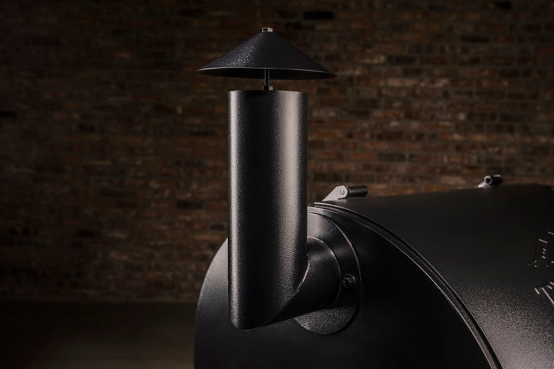 Nærbilde av en svart metallskorstein med regnhette på en Traeger Pro D2 575 pelletsgrill fra Traeger, foran en uskarp murveggbakgrunn.