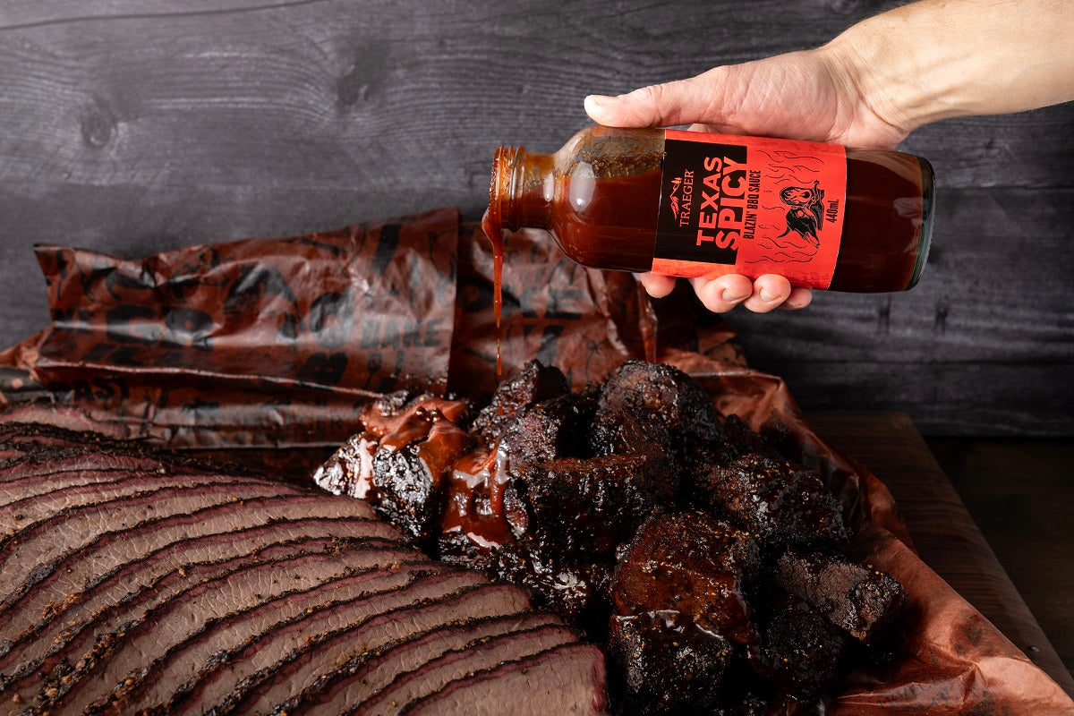 En hånd heller Traeger Texas Spicy BBQ Sauce 440 ml på røkt bryst og burnt ends plassert på slakterpapir.