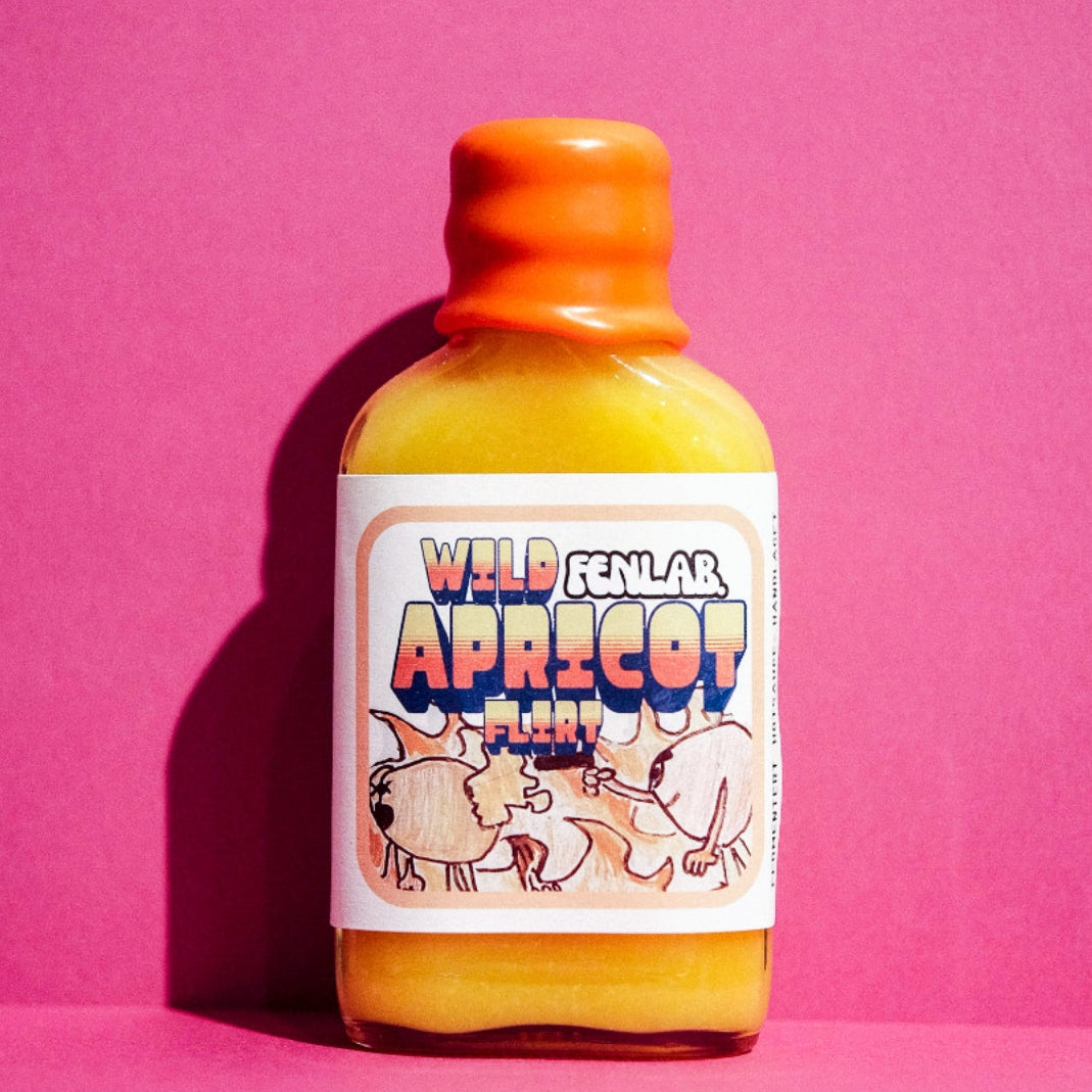 En liten flaske med oransje hette og en etikett med teksten Wild Apricot Flirt | Fen Lab hot sauce mot en ensfarget rosa bakgrunn.