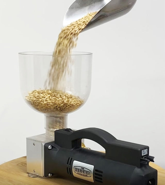 En øse øser korn i Grainfather Electric Grain Mill - Maltmølle (Uten base) fra Grainfather, som har justerbare valser og er plassert på en treoverflate med hvit bakgrunn.