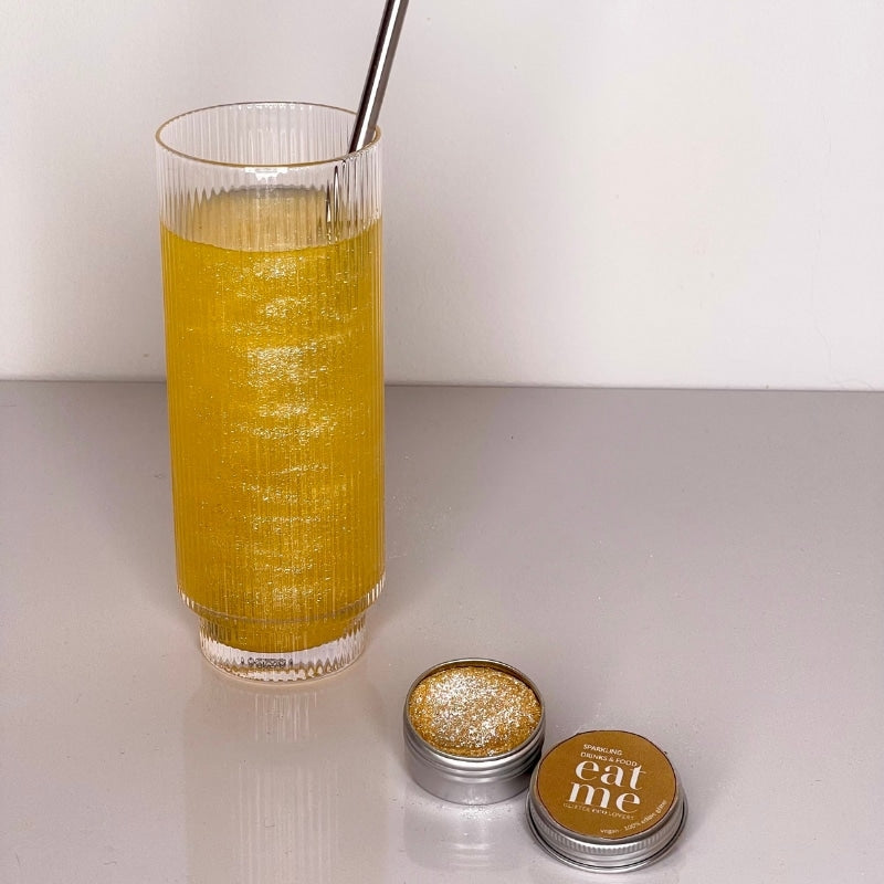 Et høyt glass med gul, glitrende drikke med metallsugerør står ved siden av en åpen Eco Lovers Eat Me Glitter Gold 150 ml boks, som inneholder spiselig glitter og spiselig måkeglitter.