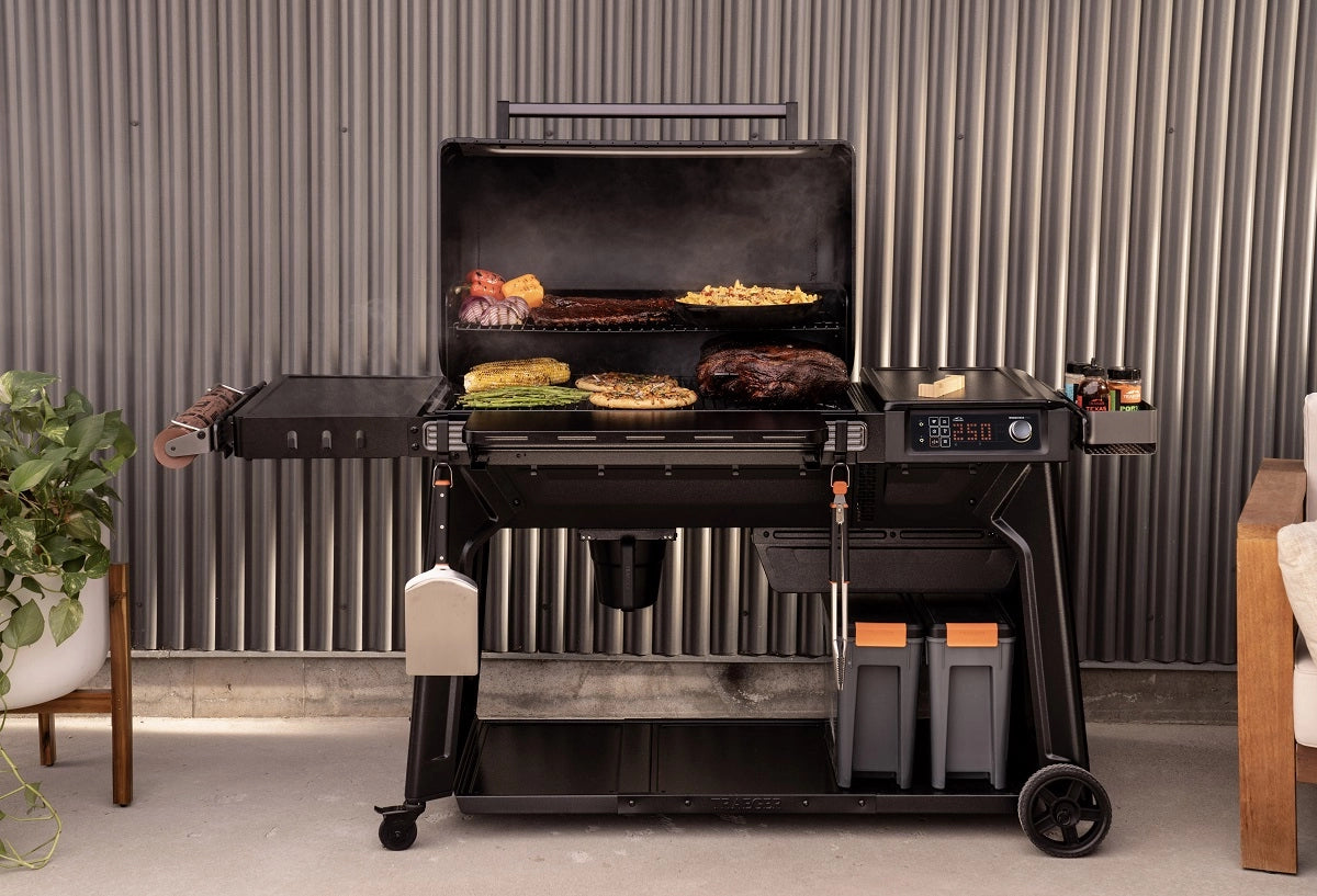 Traeger Woodridge Pro Pelletsgrill, som her vises med åpent lokk og griller grønnsaker, ribbe, mais og pommes frites, har grillverktøy og oppbevaringsbokser under. WiFIRE-teknologi gjør det mulig å grille smart utendørs.
