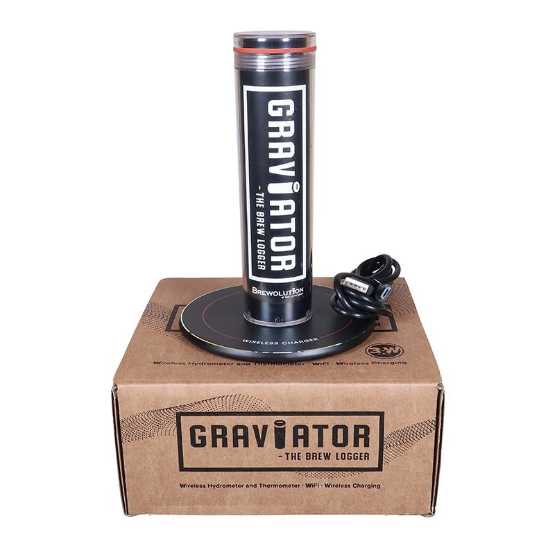 En sylinderformet Brewolution Graviator - WiFi hydrometer og termometer står oppreist på en pappeske merket "Graviator - The Brew Logger", sammen med en trådløs lader og en ladebase.