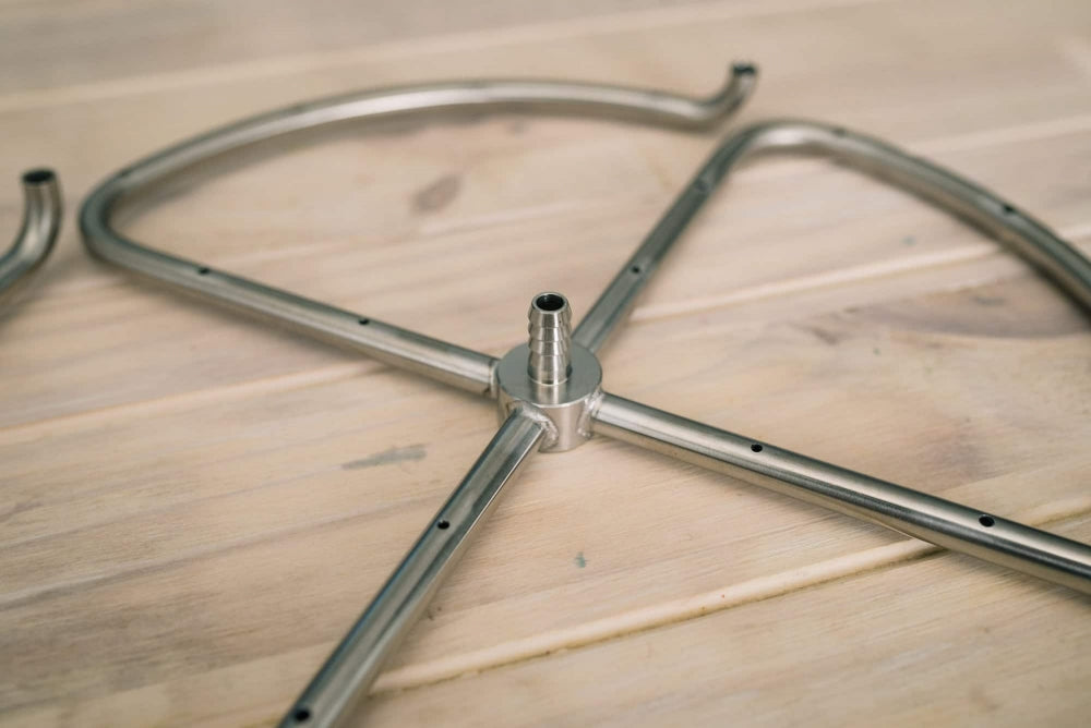 Et nærbilde av Ss Brewtech Manifold | Mash Re-Circulation for kjeler opp til 75 liter, laget av 304 rustfritt stål, viser glatte sveiser mens den hviler på en overflate av tre.