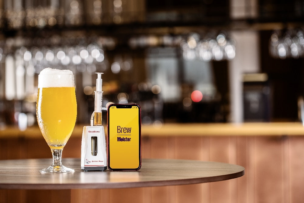 Et glass øl, en BrewPen-enhet og en smarttelefon med Brew Meister står på et barbord og viser Anton Paars EasyDens Digitalt Hydrometer for måling av konsentrasjon i drikkevareindustrien for øl og vin.
