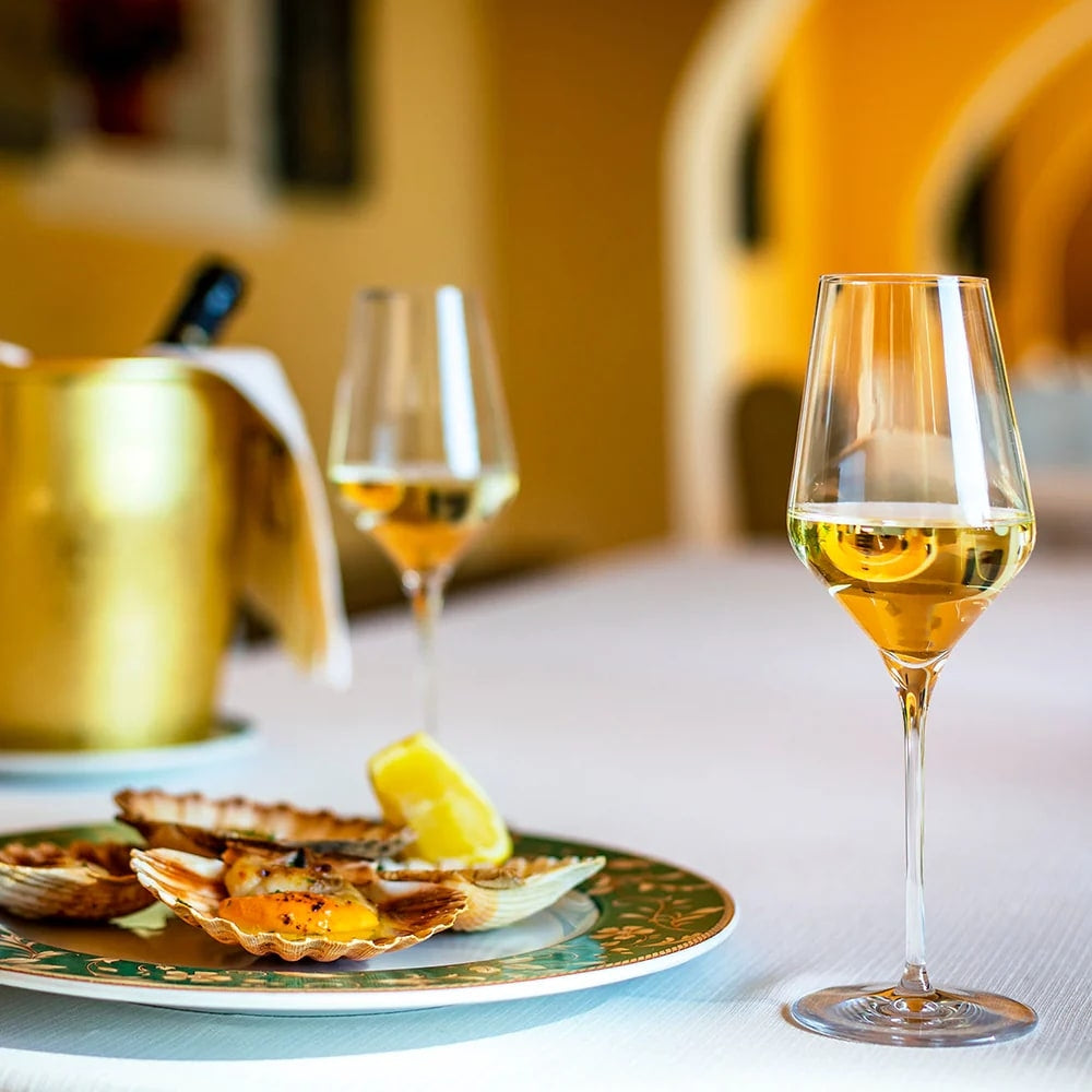 En tallerken med bakte kamskjell med sitron står ved siden av to Stölzle Lausits QUATROPHIL hvitvinsglass 405 ml og en vinkjøler i gull på bordet.