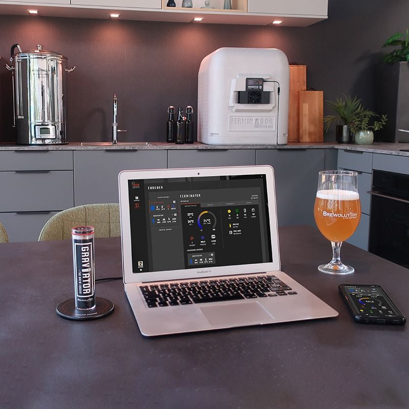 På kjøkkenbenken står en bærbar PC med BrewCreator ved siden av et glass øl, en mobiltelefon på trådløs lader, en boks og bryggeutstyr, inkludert Brewolution Graviator - WiFi-hydrometer og termometer.