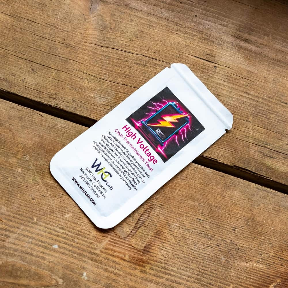På en treflate ligger en liten, hvit pakke merket "High Voltage - Electrifyingly Clean Thermotolerant Yeast 11g (kveik)" fra WHC Lab, med en lyngrafikk og produktdetaljer.