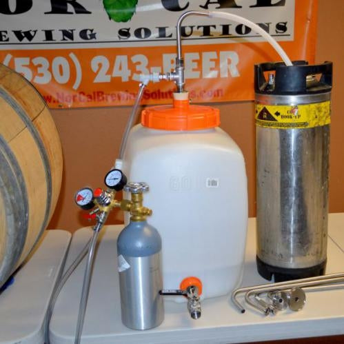 Et hjemmebryggingsoppsett med Norcal Speidel Ball Lock Transfer Tool, CO2-tank, trykkregulator, Speidel-gjæringskjele, tappestokk i rustfritt stål, metallfat og tilkoblingsslanger på et bord.